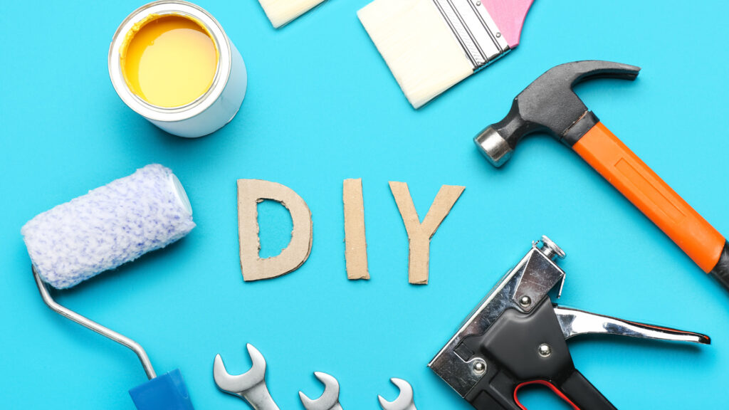 Flat lay of DIY tools, including paint, roller, hammer, and cardboard letters on a bright blue background