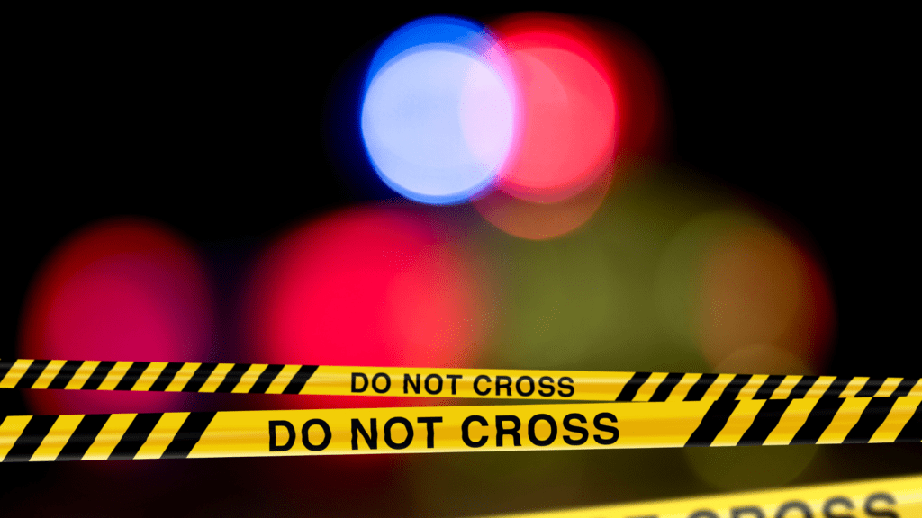 A 'DO NOT CROSS' police tape in the foreground with a blurred background of police emergency lights in red and blue, indicating a restricted area typically seen at crime scenes or emergency situations.