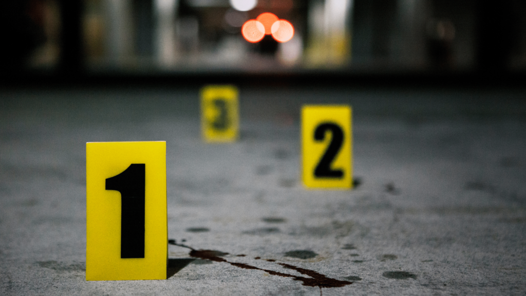 A close-up image of a crime scene with numbered yellow evidence markers '1', '2', and '3' on an asphalt surface.
