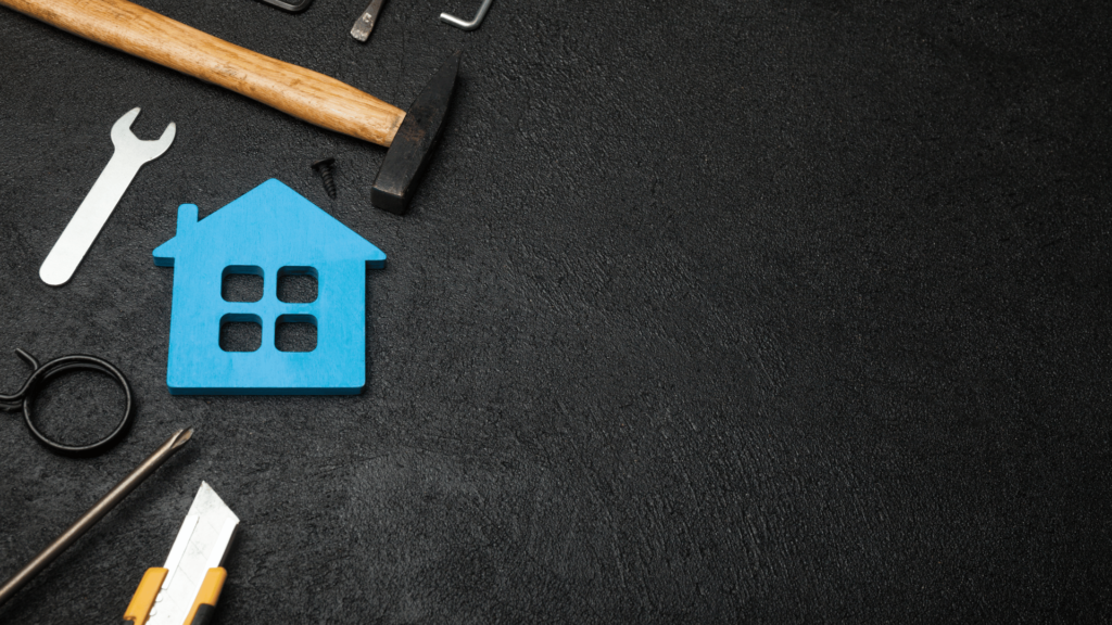Top view of a bright blue house-shaped cutout surrounded by various tools on a dark textured surface, suggesting home repair or renovation.