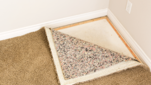 A corner of a room showing a beige carpet partially peeled back to reveal an older, multi-colored carpet underneath, with the bare floor and baseboards visible at the edges. The image depicts the process of changing or removing carpet in a room.